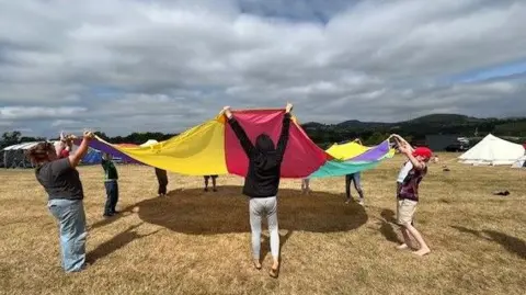 A group of adults and children stand in a circle holding a multi-coloured parachute up at head height. They are stood on dry grass which is a yellowy colour. The sky is blue but very cloudy and there is a white tent to the left of the image. 
