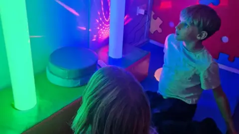 Naiomi Flewers A boy with fair hair, wearing a white t-shirt sits on the floor and looks up at fluorescent tubed lighting. A girl sits with him, with the back of her head visible, looking at the lighting. The lighting is green and there is another tube next to that which isnt lit. There are soft play toys and there is also a red laser light seen in the image. 