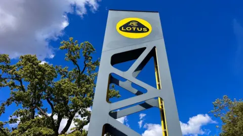 Martin Giles/BBC A tall grey sign outside the Lotus headquarters in Hethel, near Wymondham, in Norfolk. The sign has a Lotus logon which is yellow with a black curved triangle inside which says LOTUS in yellow writing. There are green trees surrounding the sign and the sky is blue as the photograph has been taken during the day.