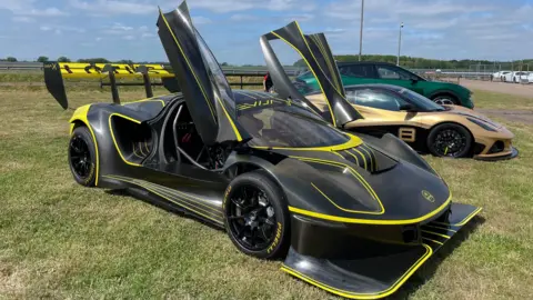 Edd Smith/BBC A row of three Lotus sports cars lined up at Lotus headquarters in Hethel, Norfolk. The cars are black and gold. 