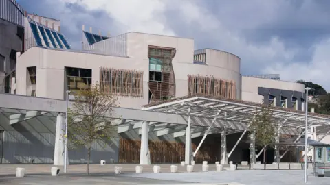 Getty Images The front of the holyrood building on a cloudy day, with white stone walls wooden slatted windows and small trees.