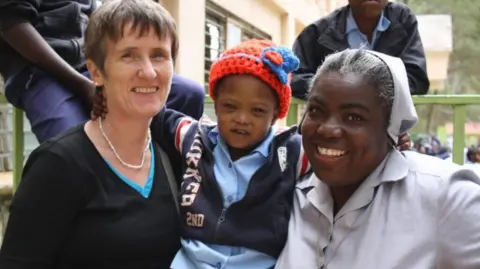 NPH International Gena, left, is smiling at the camera. She has short brown hair and is wearing a black long sleeve top. A child is in the middle, wearing a bright red hat and a navy zip up jacket. Another woman, right, is smiling at the camera. She is wearing a light grey dress and has a matching hair scarf on.