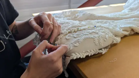 In this picture a person is sewing a wedding dress. The dress is on a table and the hands are pictured altering the gown.