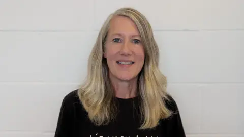 Rhiannon has long blonde hair and wears a black t-shirt. She's smiling at the camera against a white brick wall. 