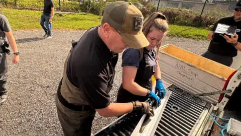 A man and a woman are measuring an eel on a silver ruler. The man is wearing green dungarees over a black T-shirt and is also wearing a green cap, the woman has her light brown hair pulled up and is wearing black dungarees over a blue T-shirt. They are working outside and there are other people standing around them.