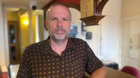 Huw is looking at the camera, he has a grey goatee and dark brown eyes. It is a head and shoulders shot of him stood inside a building which is blurred behind him. He is wearing a brown shirt with a dark yellow and green circular pattern on it.