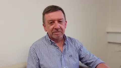 Mark Farrell. He has short dark brown hair and light grey beard, wearing a blue and white striped shirt.