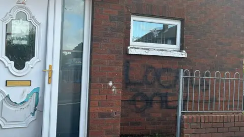 The words "Locals only" are daubed on the side of a house in black paint under a small window. The front door is damaged.
