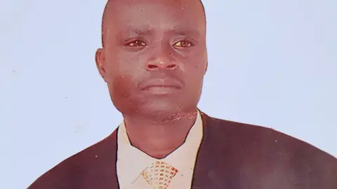 A photograph of a snapshot of Samuel Owino Owoyo. He looks serious in a dark suit jacket, white shirt and spotted tie.
