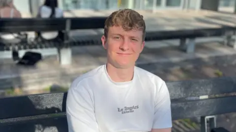 Oliver Reaper, 24, who sits on a bench in Central Square, Cardiff, outside the bus station. He has short curly blond hair and he holds his left arm with his right hand while wearing a white t-shirt which reads 'Los Angeles' in Gothic script. The image focuses on him but behind him there are a couple of women on a bench, the background is blurred.