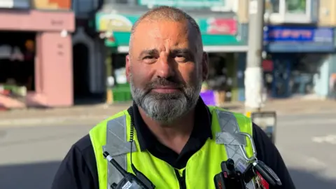 Dave, a man with a shaven head and grey beard wearing a black shirt and a yellow hi-vis vest looks directly into the camera. He is also wearing a radio with an earpiece and a body worn camera. He is standing near a sunny high street.
