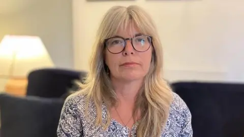 BBC Liz Hopkin looks directly at the camera. She wears round black lense reading glasses and has blue eyes and blonde hair. She wears a vest top and a paisley jacket. A blurred living room lit up by a side table lamp can be seen behind her in the background. 