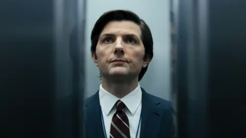Getty Images The actor Adam Scott standing in an elevator as the doors close. Between the closing doors we can see he is wearing a sharp suit and tie. He is clean shaven and has well-kept black hair.