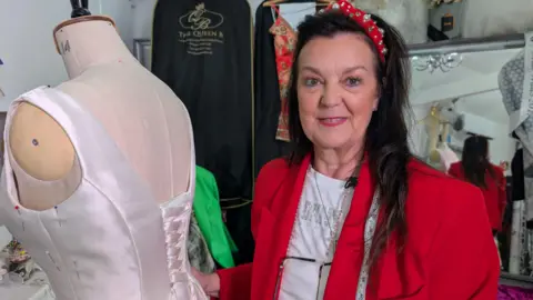 In this picture Angie is smiling at the camera as she stands next to a mannequin. Angie wears a red blazer and a white t-shirt.  She's also wearing a sparkly red headband and has long black hair. 