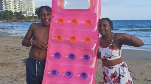 Family Ameiya (R) and Ricardo Del Brocco (L) on holiday. They are either side of an pink inflatable. They are on a beach and smiling. Ameiya has short dark hair and wears a two piece costume of white with a floral design. Ricardo has short dark hair in braids and wears a blue pair of shorts.