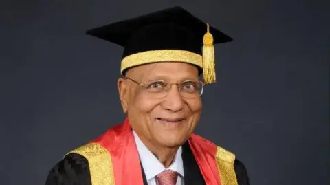 University of Wolverhampton A man in a gradation cap and gown smiles at the camera. His gown has a red and gold hood lining and his cap is also lined with gold fabric.