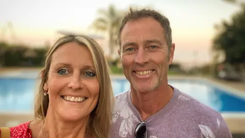 Family handout A man and woman smile on holiday. The woman has shoulder length blonde hair and is wearing a red top, the man has short brown hair, stubble and a lilac t-shirt. There is a pool behind them