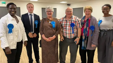 BBC A group of politicians in a line, wearing smart clothing, and smiling for the picture after a by-election.