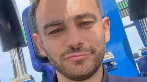 Family handout Corrie Lee Stavers takes a selfie. He is a young man with short dark hair and neat thin dark beard. He is wearing a navy polo shirt and appears to be in the seat of a blue funfair ride.
