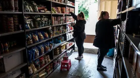 Getty Images Two workers in a food bank organise supplies on shelves, which have supplies of tinned and dry food. There is a step on the floor to reach the higher shelves and a window behind the workers.