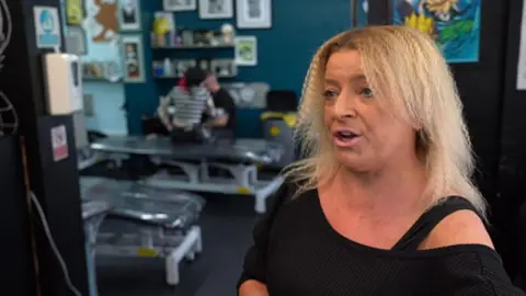 BBC A middle-aged woman with shoulder-length light blonde hair. She wears a black shirt and stands in a tattoo studio with several tattoo beds behind her.