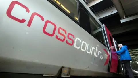 PA Media Taken from a low angle near the floor, the photo shows the side of a grey train, with "Crosscountry" written on it in large text - cross in red, country in yellow. A man in a blue coat is getting onto the train in the far corner of the image.