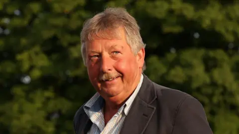 PA Media Sammy Wilson has greying hair and a grey moustache and is wearing an open-necked light striped shirt and a dark grey jacket  