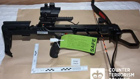 West Midlands Counter Terrorism Police A black crossbow with a right-angle ruler beneath and a green police label attached reading "Safe" and other details that cannot be made out. The weapon is sitting on light brown paper