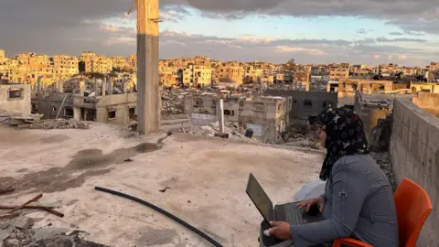 Handout Manar sits on an orange chair with a laptop on her lap. She is looking out into the distance and wearing a dark blue jacket. In the background are buildings falling apart and with roofs off and various bits of rubble.