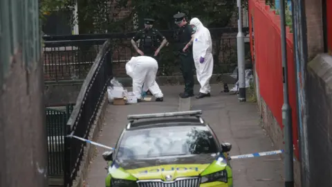 PACEMAKER A PSNI car is parked in front of a walkway. Two PSNI officers are standing next to two people in forensic suits, one is crouched down sorting through carboard boxes and plastic bags.