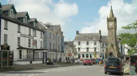 BBC A main street in Machynlleth leading to a tall stone clock tower in a square and townhouses around the edges. There are cars on the road. It is a blue sky with some clouds.