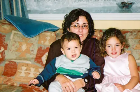 Sarah Ezekiel A family photo taken in the 90s of Sarah and her two children. They are sitting on a patterned sofa with autumn colours. Sarah has her toddler son on her lap and her daughter sitting next to her.