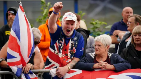 PA Media A group of people are standing at a metal barrier. A woman wearing sunglasses holds a Union flag. There is a man in the middle of the scene wearing a waistcoat in Union flag colours. The waistcoat is covered in pin badges. The man, who is also wearing a cap, has an arm raised as he shouts. A woman next to him is looking into the distance while leaning on a flag draped over the barrier.