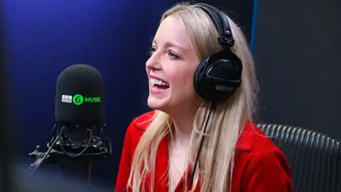 BBC Lauren Laverne laughing into a BBC Radio 6 Music microphone. She has long, blonde hair and is wearing large black headphones and a red shirt.