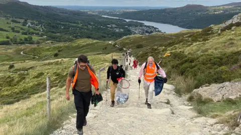 BBC Three people walking up a steep rocky mountain path with a lake in the background. Two men, left and right, are wearing orange tabards and a woman is in a black top.  All three are carrying rubbish bags and the woman and the man on the right are also carrying litter picking grab sticks.