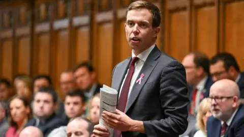 House of Commons/Reuters A man in a grey suit with red tie stands in the House of Commons to ask a question. He is holding some folded papers. 