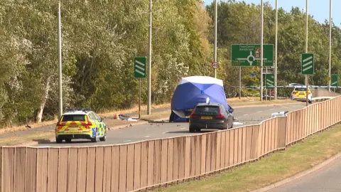 BBC A police cordon at the scene. The road is closed in both directions. A tent covers the wreckage of a car.