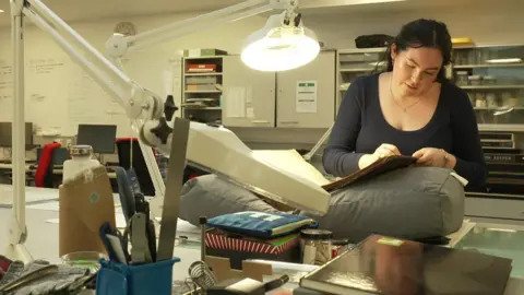 BBC A woman in an office looking through an old ledger with an overhead light over it.