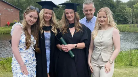 Edge Hill University Stephanie Kennedy and daughter Mia both have shoulder length blonde hair and are wearing black mortar boards and academic gowns. He other two daughters also have long blonde hair - one is wearing white dress with blue flowers and the other wears fawn trousers and matching sleeveless top. Her husband in the background has greying and is wearing a sky blue Oxford shirt