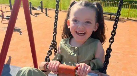 BBC Niamh is photographed on a swing in playpark. She has her hair in two pigtails at the sides, held in place by pink bows. She is smiling. She has blue eyes and is wearing a green top.