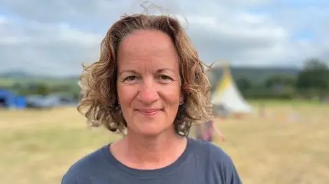 BBC A woman with light brown wavy hair, wearing a navy crew neck t-short smiles as she looks into the camera. She is standing in a campsite with tents and hills obscured in the background. The background is blurred and it is a head and shoulders shot of her. 
