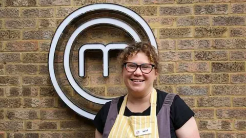 BBC Beth Hodgson, who has curly brown hair, stands in front of the brick wall with a white MasterChef logo fixed to it. She is wearing a yellow and white pinstriped apron with her name shown on a tag. Beneath the apron she is also wearing a black T-shirt and navy overalls.