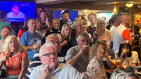 BBC Inside the Colliers pub in Tyldesley packed with friends and family drinking and watching intently the late Tim McCarthy on the Chase. Tim McCarthy is shown on a TV screen in the background taking part in the show.