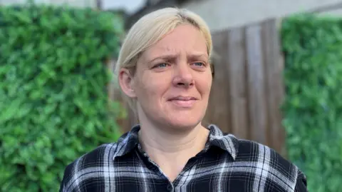 BBC Picture of Jen in her back garden. There is a fence and bushes behind her. She has blonde hair, scraped back behind her ears. She is wearing a black and white checked shirt.