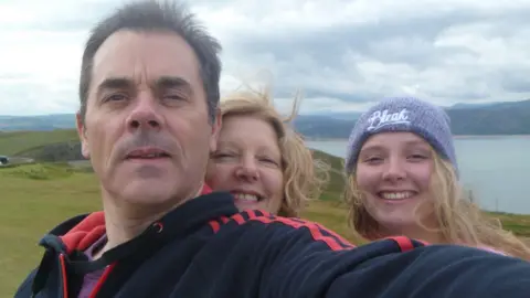 Family A picture of a man, woman and a teenage girl smiling at the camera standing on top of a hill. Mountains and the sea are in the background.