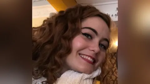 Suffolk Police A smiling Courtney Mitchell looks directly at the camera with her head tilted to her left. She is a young woman with long, slightly curly dark brown/ginger hair and is wearing a white scarf.
