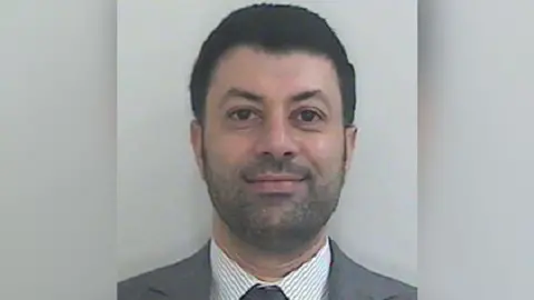 HMRC Image shows a Passport-style photo of Arif Patel, who has short dark hair, a short dark beard, and is wearing a grey suit with a grey and white striped shirt and grey tie.