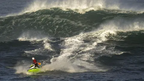 Adrian Bagley A RNLI lifeguard riding a jetski through a large set of waves. The jet ski is lime green, the lifeguard is wearing a white safety helmet and a red life jacket. He appears small compared to the size of the waves.