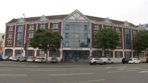 BBC The outside of a large building. The centre is a large window the full length of the building with many windows across it. The frames are blue and white and it is red brick. A number of cars, mostly white and silver, are parked outside facing towards the building. A Salvation Army logo can be seen on one of the windows in the centre of the building, above a sign reading "Centenary House"
