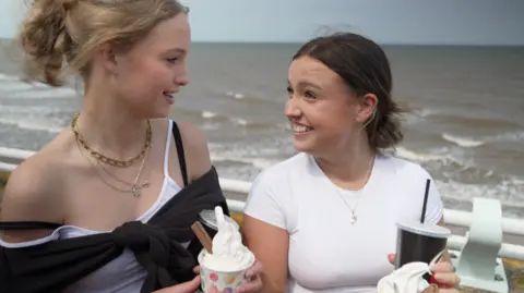 BBC Two teenage girls turn to smile at one another. They are both holding Mr Whippy-style ice creams with chocolate flakes, in tubs. The sea is not far behind them.